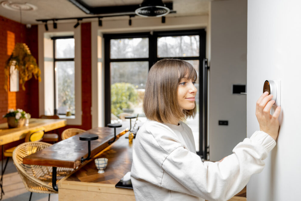 Modern kitchen, smart thermostat focus, wide shot with wooden furniture and large windows, bright daylight, editorial travel photo, one woman.