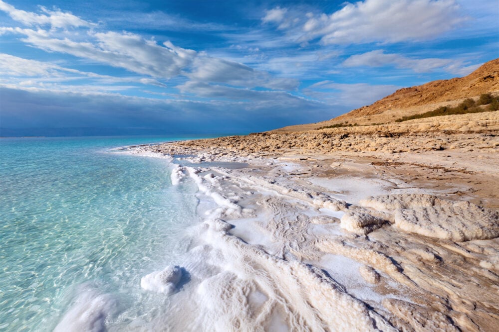 Dead Sea shoreline, salt-encrusted rocks, turquoise shallows meeting beach, midday sunlight, editorial travel photo, no people.