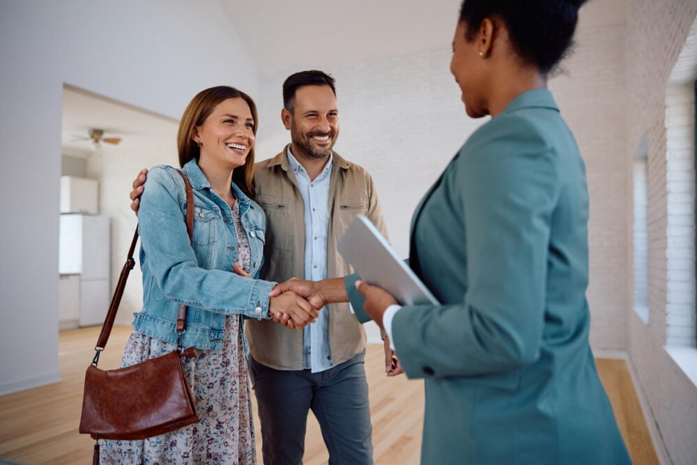 Bright empty room, handshake in focus, group posed front-center, daytime natural light, editorial travel photo, three people.