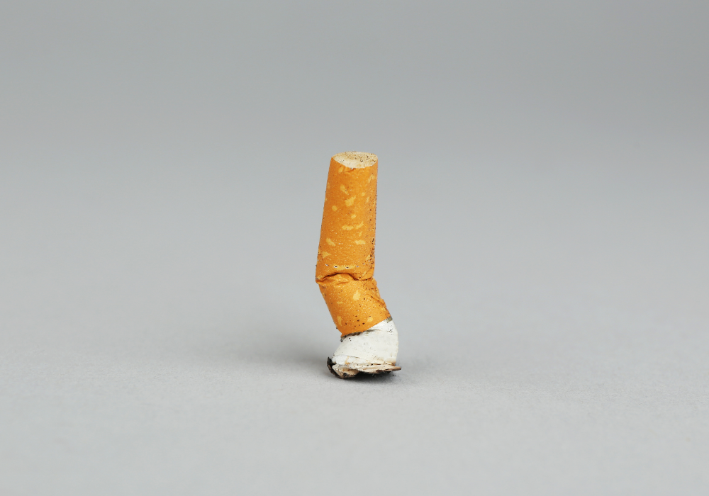 Smooth gray tabletop, upright cigarette butt center, side lighting, neutral palette, editorial travel photo, no people.