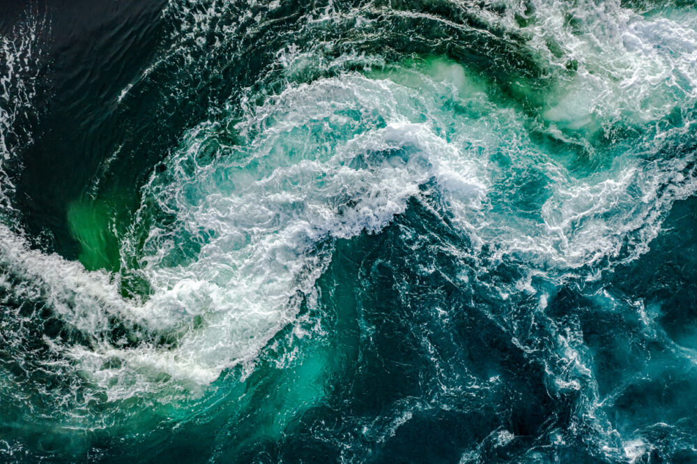 Open ocean, dramatic whirlpool center, circular wave patterns overhead, daylight highlights water hues, editorial travel photo, no people.