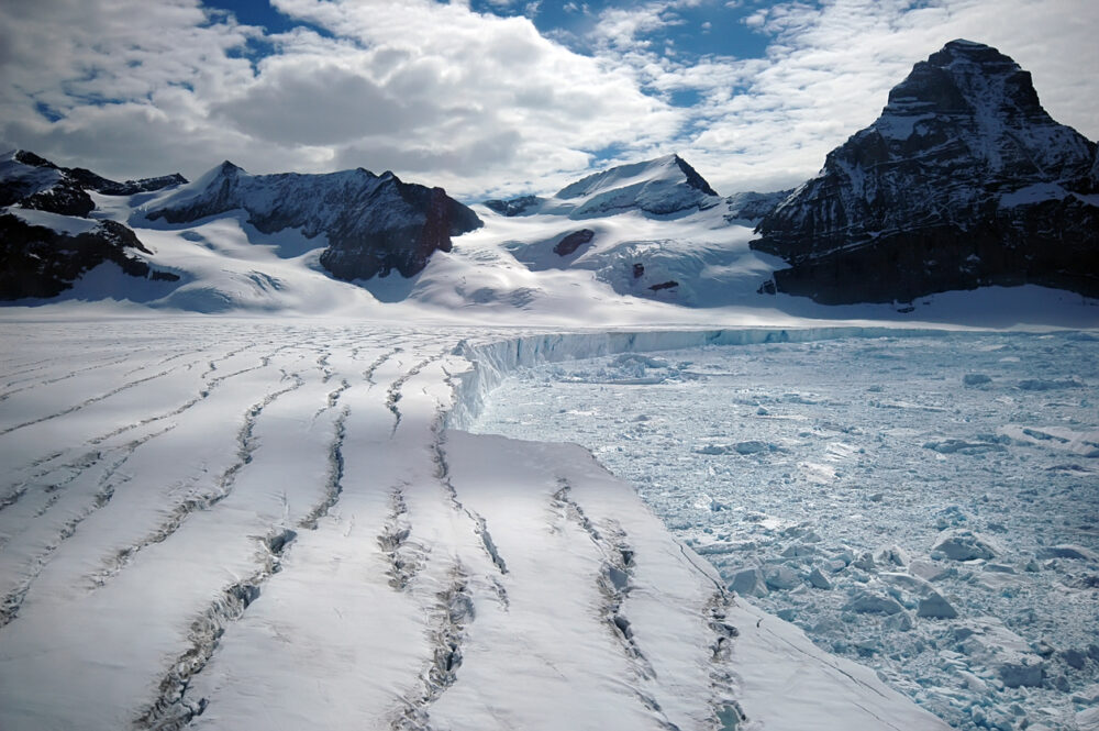 Mountain glacier, deep ice fissures foreground, fractured ice shelf midground, daylight with clouds, editorial travel photo, no people.