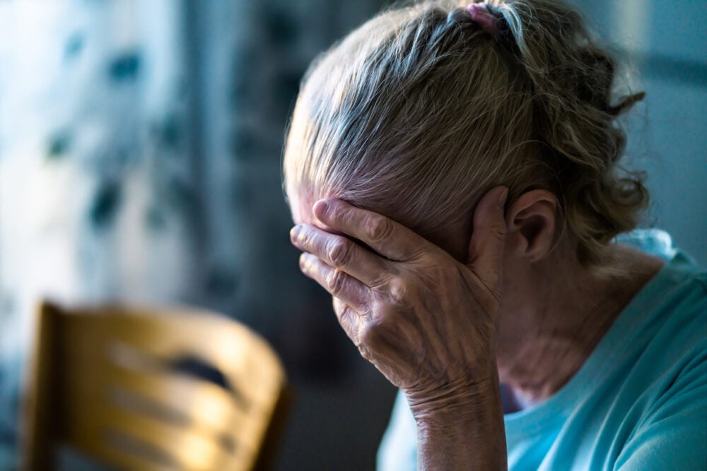 Indoors setting, distressed older woman foregrounded, midday natural light, documentary style, soft focus, people present.