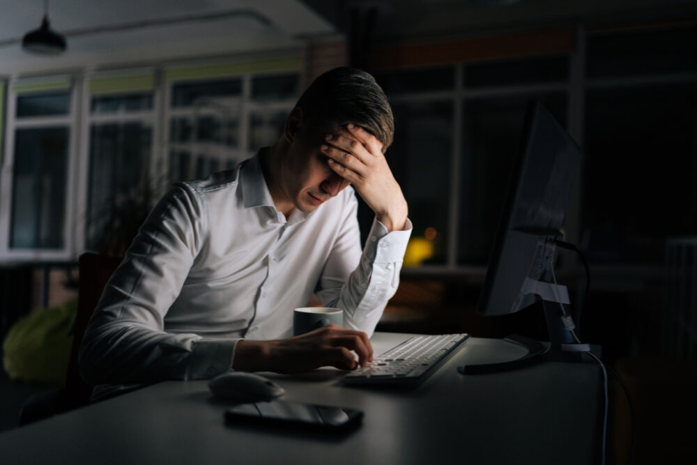 Dimly lit office, stressed man, head in hand at desk, glowing monitor at night, documentary style, people.
