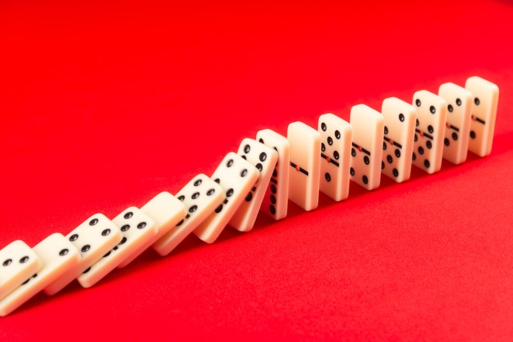 Bright red backdrop, toppling white domino row, frontal view, vivid daylight, editorial travel photo, no people.