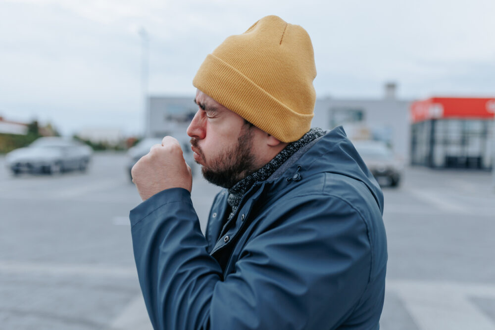 Parking lot, man coughing into fist, mid-frame, cloudy daylight, documentary style, one person.
