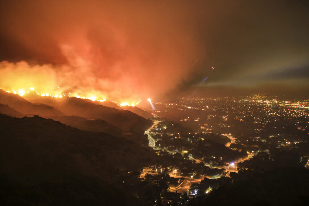Hillside wildfire, glowing flames, thick smoke rising above city lights, night scene, documentary style, no people.