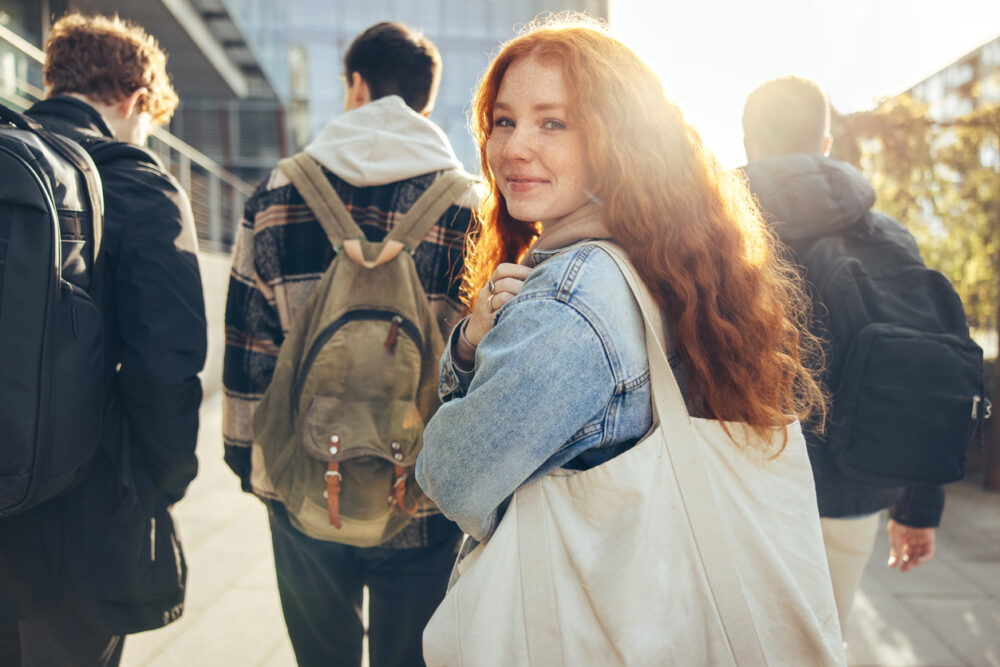 Outdoor walkway, red-haired woman in denim jacket foreground, group walking with backpacks, bright sunlight, editorial travel photo, people.