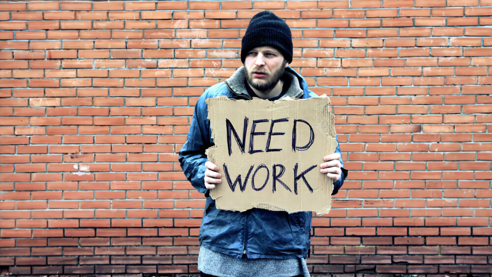 Brick wall, man with NEED WORK sign, medium shot center, overcast light, documentary style, one person visible.