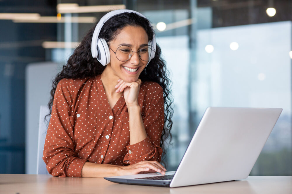 Modern office, woman in glasses and headphones at laptop, seated at desk, daylight, editorial work portrait, people.