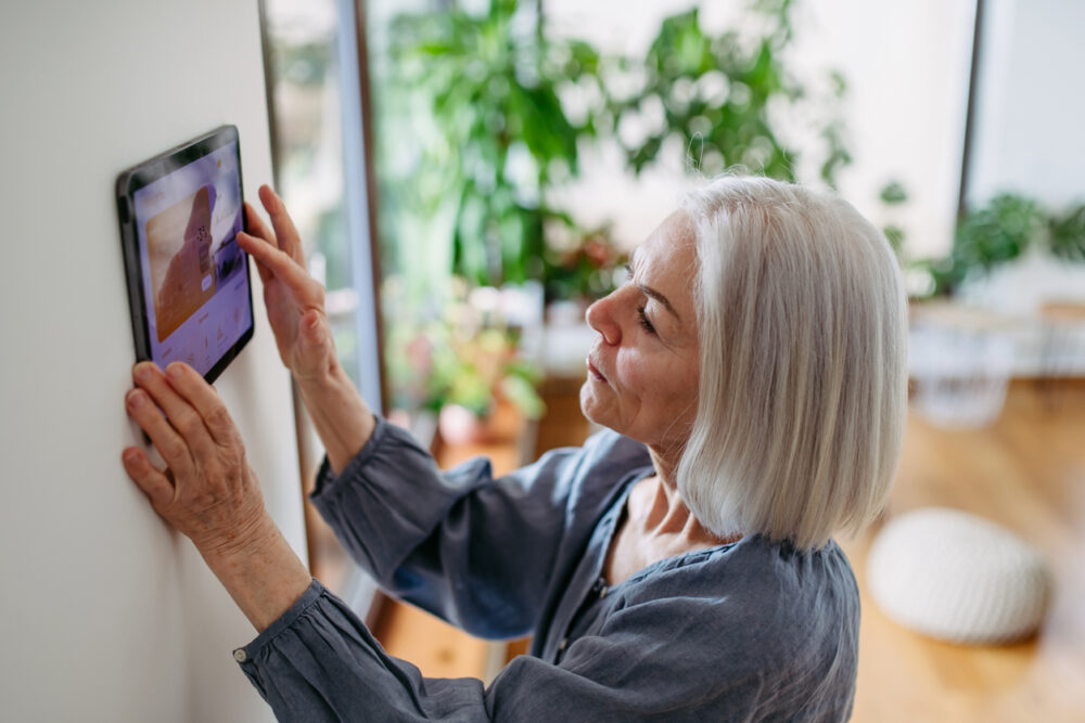 Bright sunlit room, wall-mounted tablet interaction, greenery-filled interior, morning light, editorial travel photo, older woman.