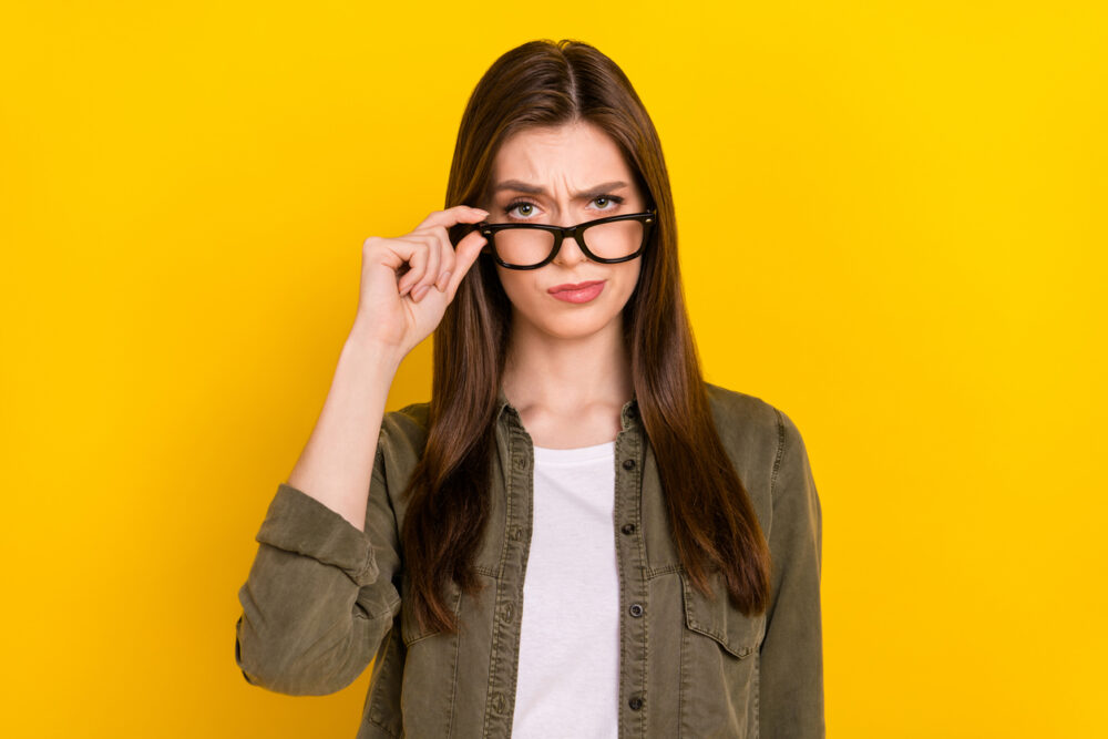 Bright yellow background, skeptical woman with brown hair and glasses, waist-up portrait, daytime lighting, editorial travel photo, one person.