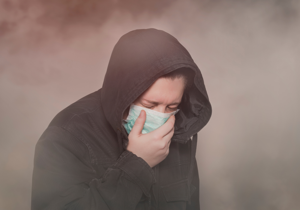 Urban street, person in hooded jacket and mask coughing, dense smoky atmosphere, overcast light, documentary style, single figure.
