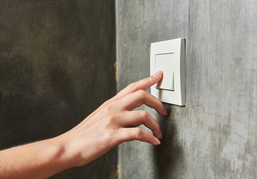 Gray concrete wall, white light switch, hand reaching in frame, daytime natural light, documentary style, one person present.
