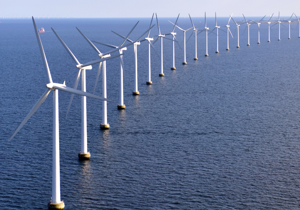 Ocean horizon, offshore wind turbines, linear formation with red ship distant, daylight clear sky, editorial travel photo, no people.