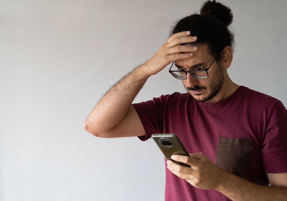 Plain light backdrop, worried man in maroon shirt, hand on head, daytime soft lighting, documentary style, one person.