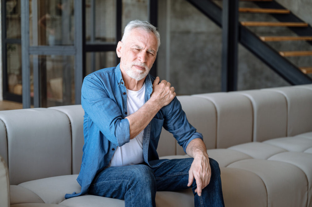 Beige couch, older man gripping left shoulder, tight composition, daytime soft light, documentary style, single person.