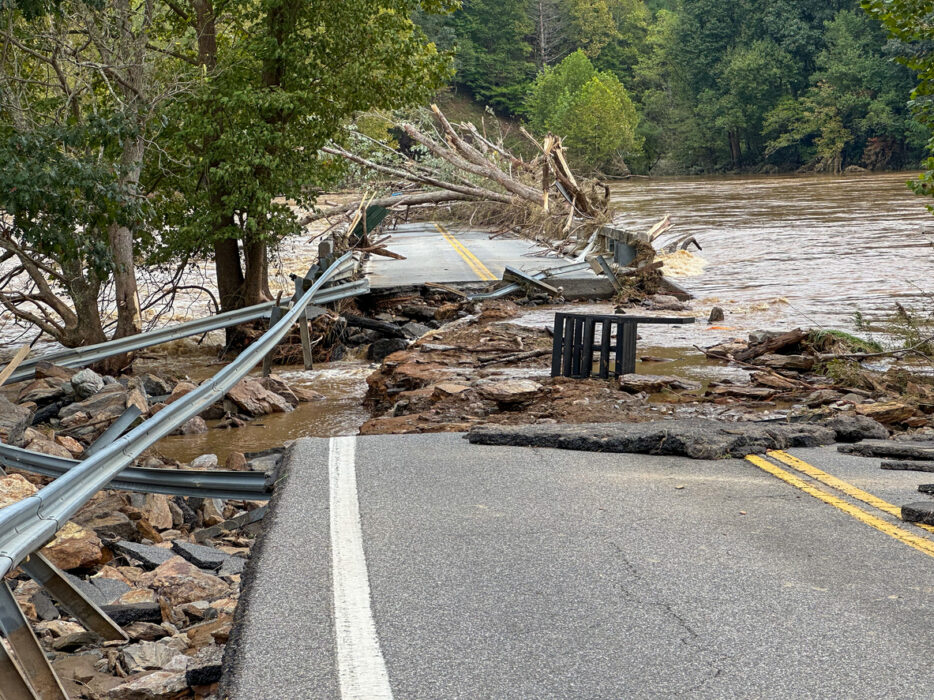 Flooded riverside road, shattered pavement and twisted guardrails, wide shot, overcast daylight, documentary style, no people.