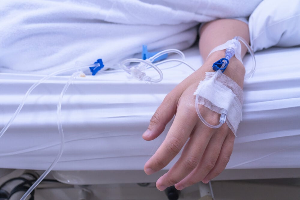 Hospital room, IV line in hand, arm resting atop bed under white sheet, soft daylight, documentary style, one person.