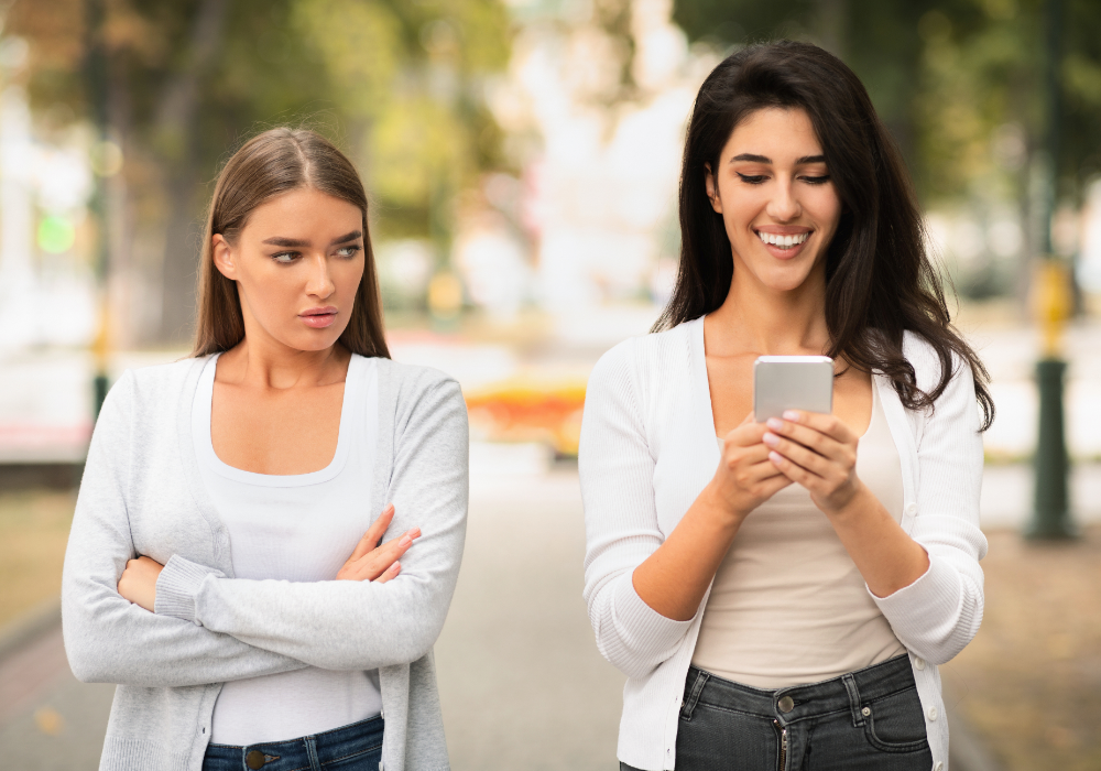 Park path, two women center frame, one smiling at phone, one arms crossed, daylight, documentary style, people present.